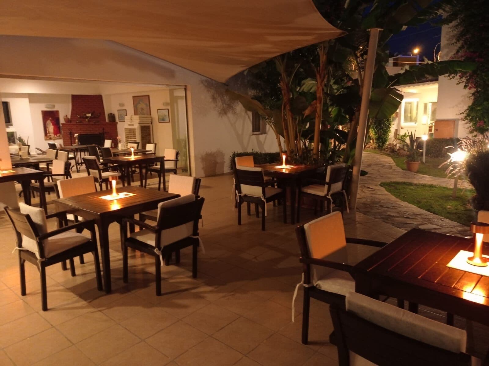 An outdoor dining area with tables and chairs, some with candles. There is a covered seating area and a view of a building in the background.