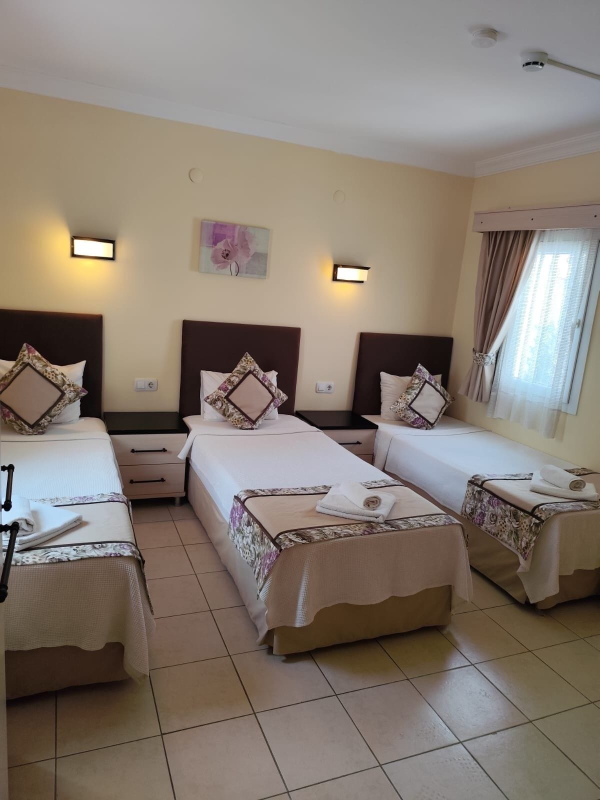 Une chambre d’hôtel avec trois lits simples, chacun doté d’une tête de lit, de tables de chevet et d’une petite table de nuit. Il y a une fenêtre avec des rideaux, un luminaire fixé au mur et un petit tableau au-dessus des lits.