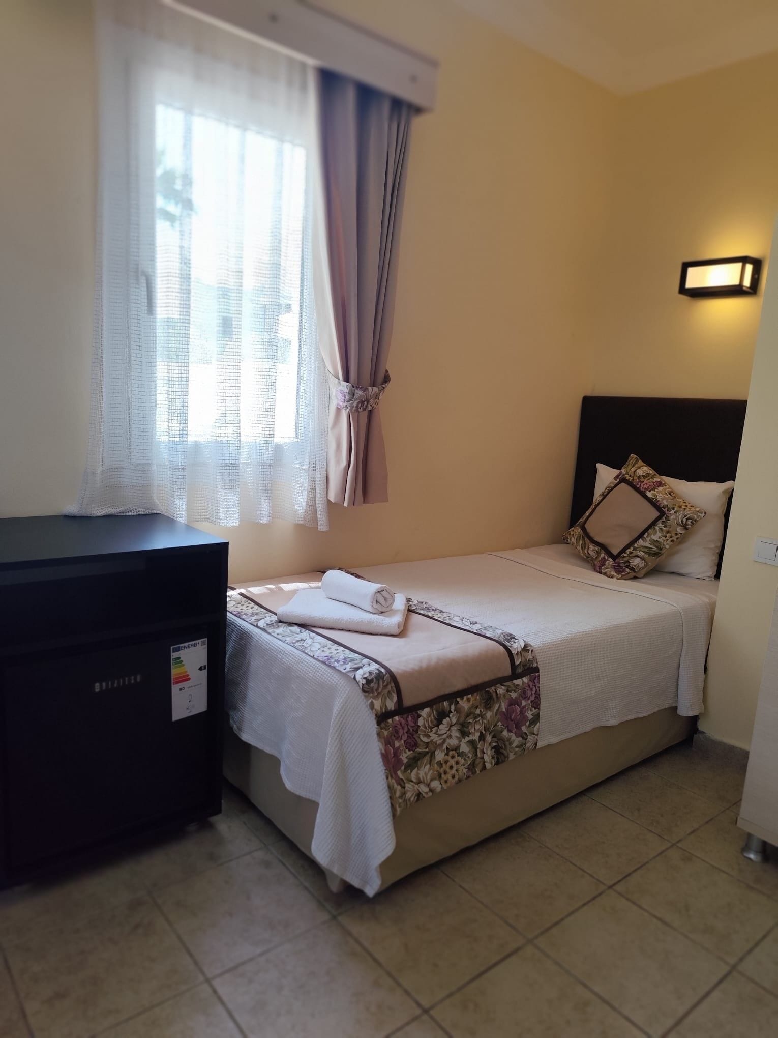 A single bed with a floral bedspread, a bedside lamp, and a rolled towel on the bed.