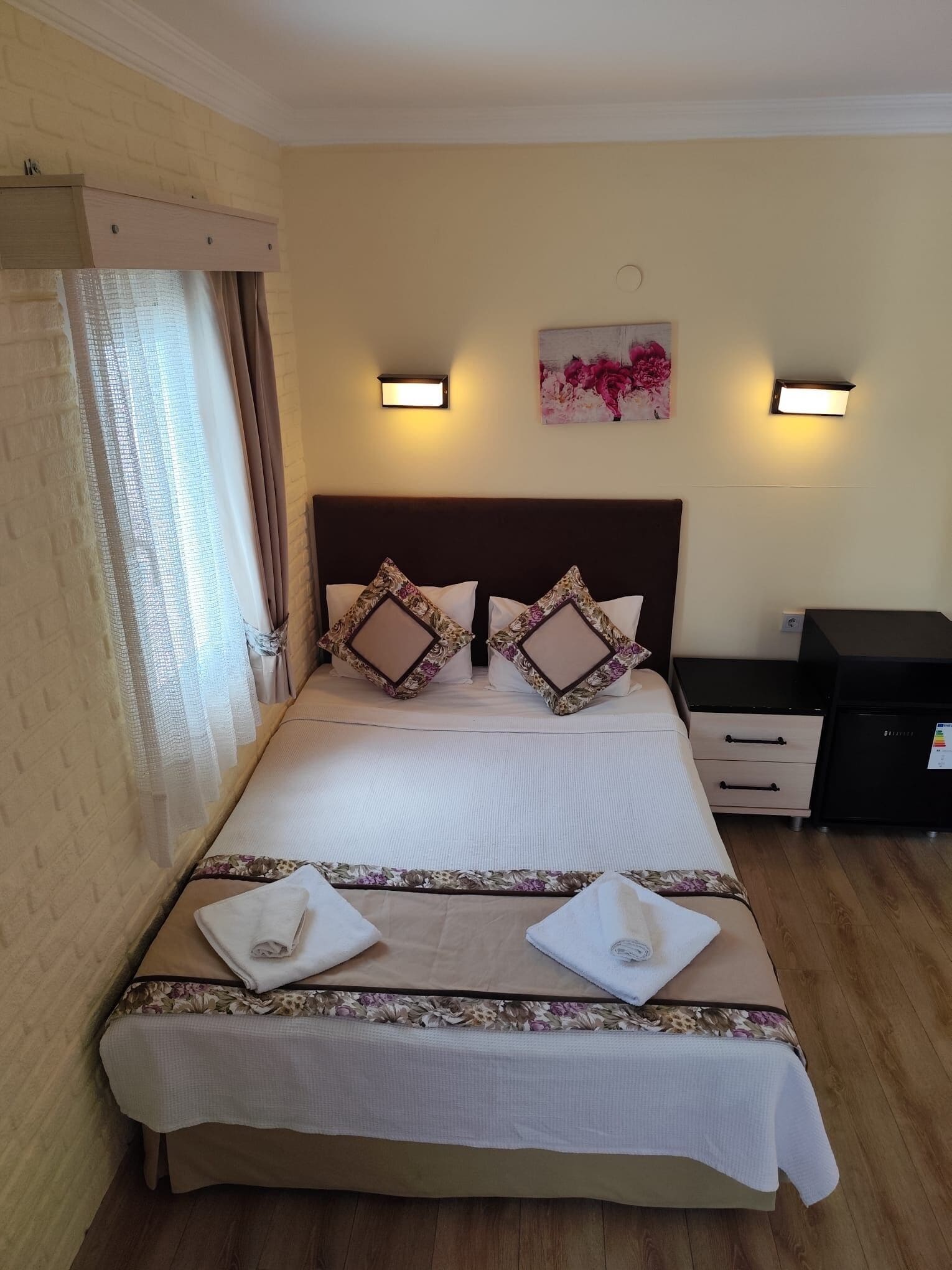 Una cama bien hecha con cabecera, dos mesitas de noche, una lámpara de pared y un cuadro con motivos florales.