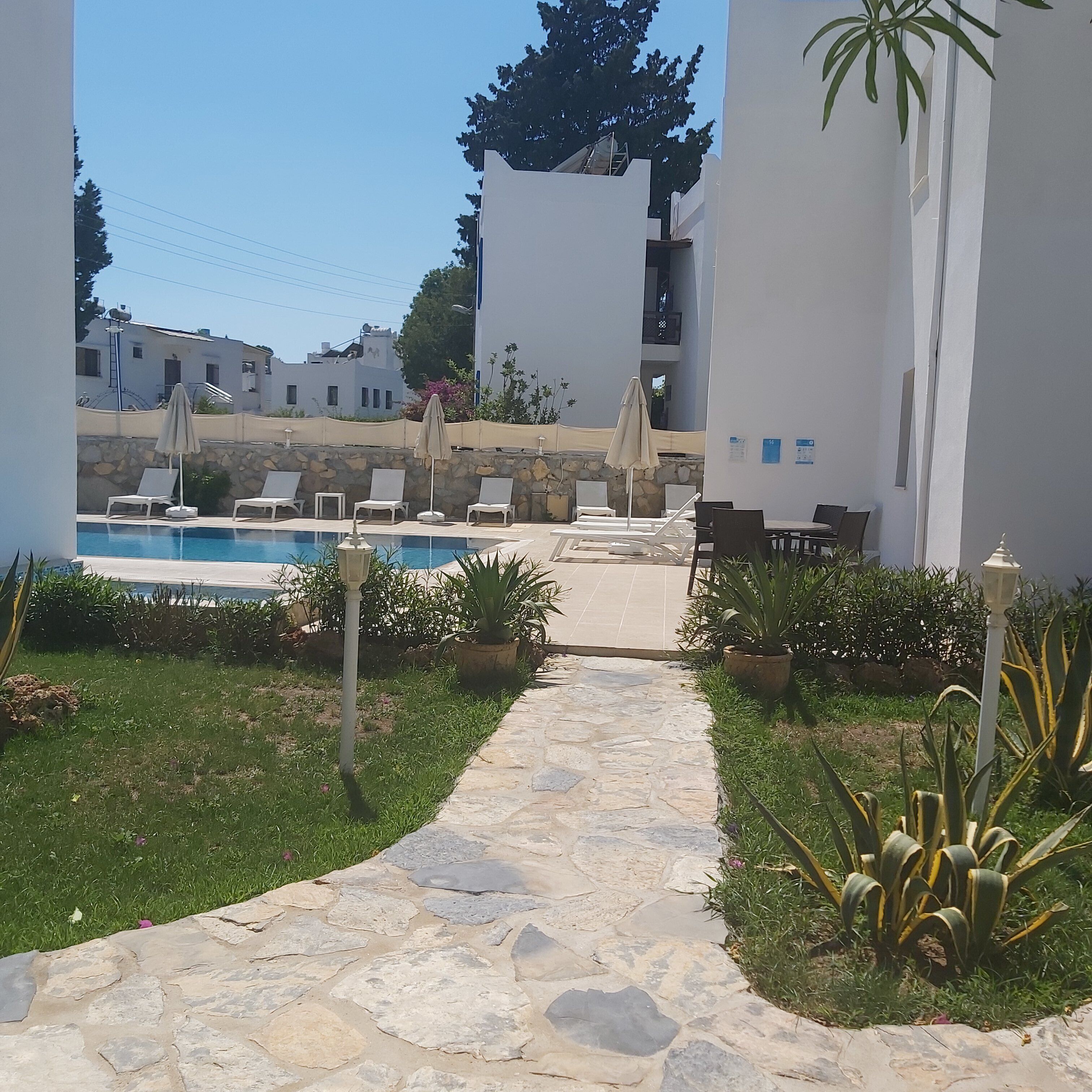 A stone pathway leading to a pool area with lounge chairs and umbrellas, surrounded by greenery and white buildings.