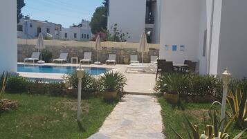 A stone pathway leading to a pool area with lounge chairs and umbrellas, surrounded by greenery and white buildings.