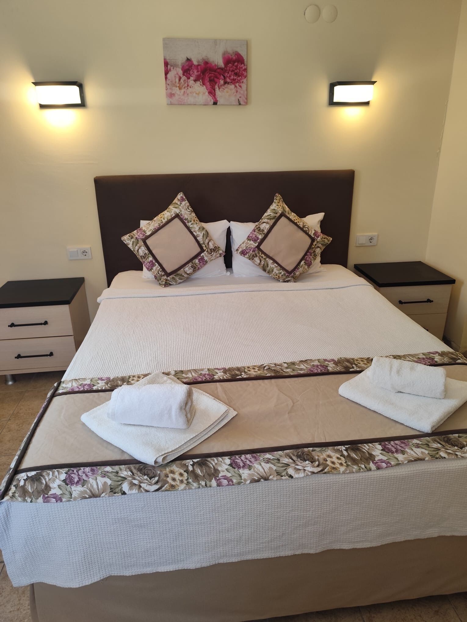 A neatly made bed with floral-patterned pillows and a matching headboard, flanked by bedside tables with lamps, in a well-lit room.