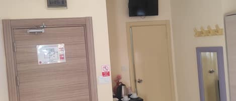 Habitación de hotel con puerta de madera, televisor montado en la pared, una mesita con una tetera y tazas, y un espejo con marco morado.