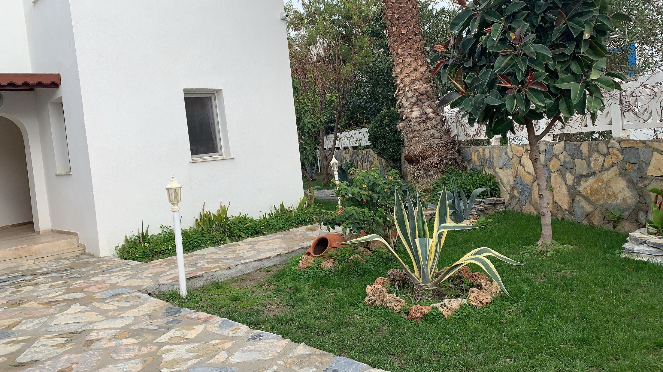 A white building with a stone pathway leading to a door. There is a small window and a lamp post in the garden area.