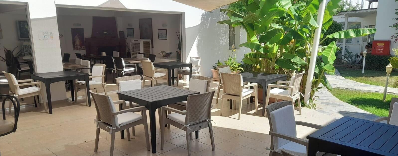 An outdoor patio area with multiple tables and chairs, surrounded by plants and a white wall.