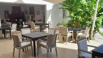 An outdoor patio area with multiple tables and chairs, surrounded by plants and a white wall.