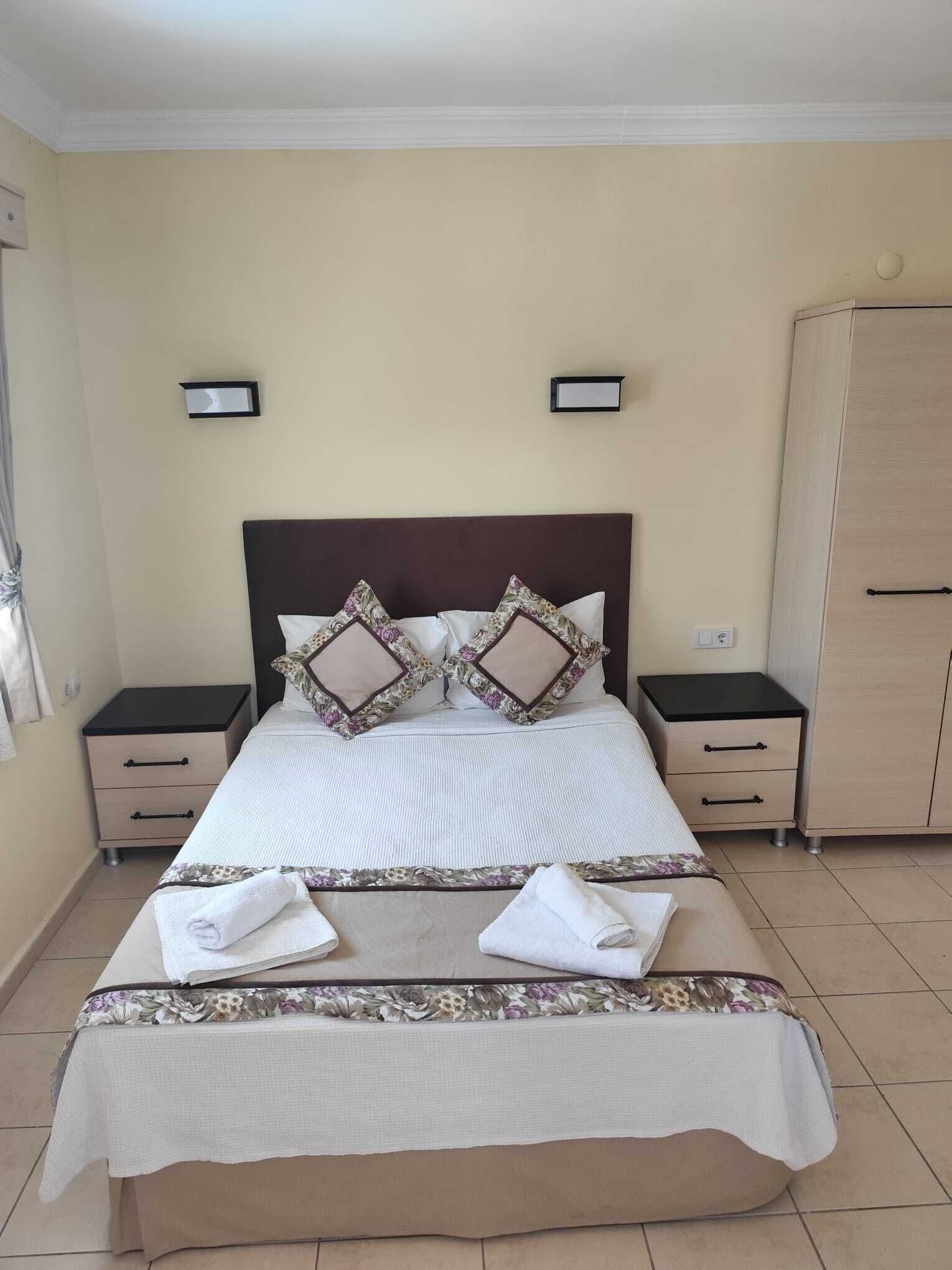 A neatly made bed with a headboard, two bedside tables with drawers, a wardrobe, and two wall-mounted light switches.