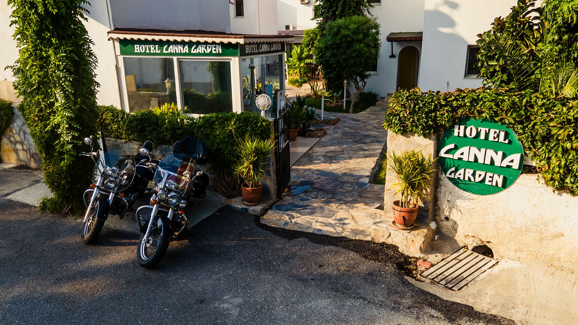 A hotel entrance with a sign reading "HOTEL CANNA GARDEN," a paved driveway, and motorcycles parked outside.
