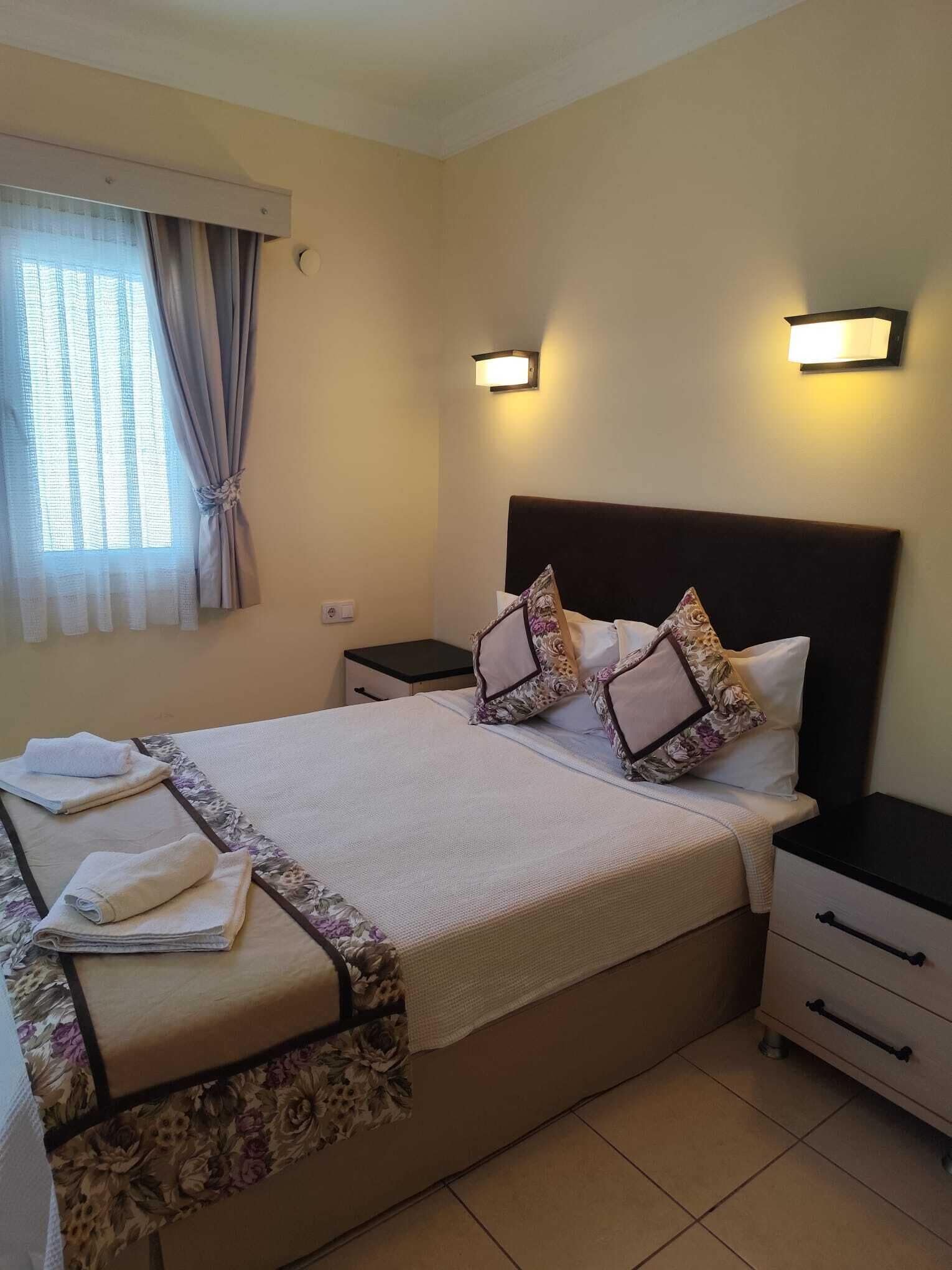 A hotel room with a bed, bedside tables, a nightstand, a window with curtains, and wall-mounted lights.
