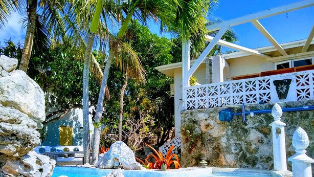 A pool surrounded by rocks and palm trees, with a white fence and a building in the background. - Ocean Views, Private Pool, and Lush Botanical Garden  (Governor's Harbour)