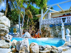 A pool surrounded by rocks and palm trees, with a white fence and a building in the background.