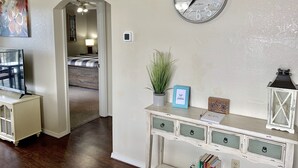 Una habitación con una cama, un televisor, un reloj, un escritorio con cajones, una planta y un cuadro enmarcado.