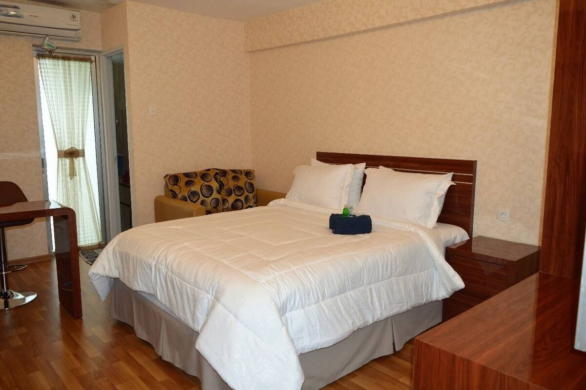 A neatly arranged bedroom with a large bed, a wooden nightstand, a small table, and a patterned armchair.