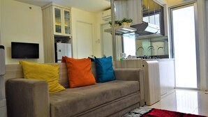 Una sala de estar con un sofá, cojines coloridos, un televisor de pantalla plana y una zona de cocina con ventanal.