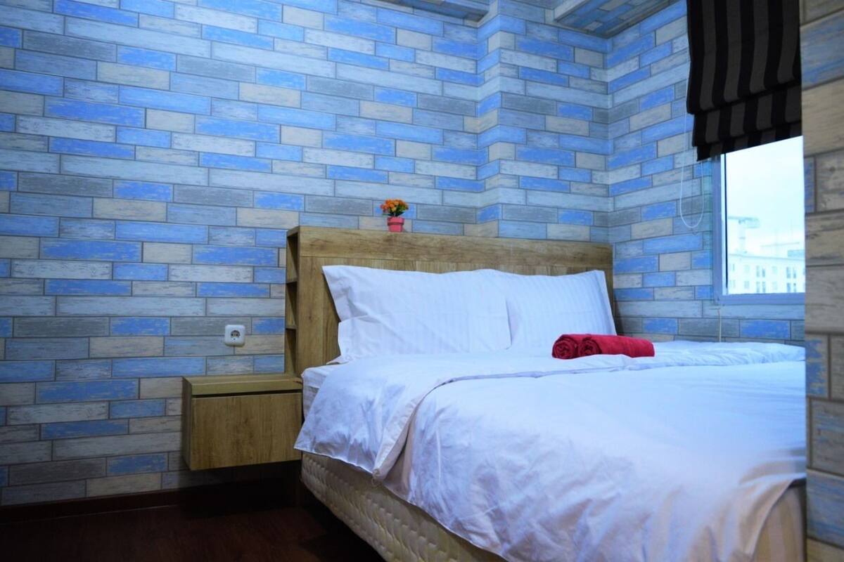 A bedroom with a brick-patterned wall, a wooden headboard, a white bed with a red towel, and a nightstand with a small plant.