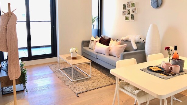 A modern living room with a grey sofa, white cushions, a coffee table, and a dining table with a plate of cookies and two mugs. - Deluxe and Brand New Lakeside Apartment (Melbourne)