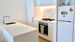 A modern kitchen with a white countertop, stainless steel sink, built-in oven, and a white refrigerator with energy rating stickers. - Deluxe and Brand New Lakeside Apartment (Melbourne)