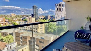 A balcony with a view of a cityscape, including modern buildings and a crane. There is a chair and a small table on the balcony. - Deluxe and Brand New Lakeside Apartment (Melbourne)
