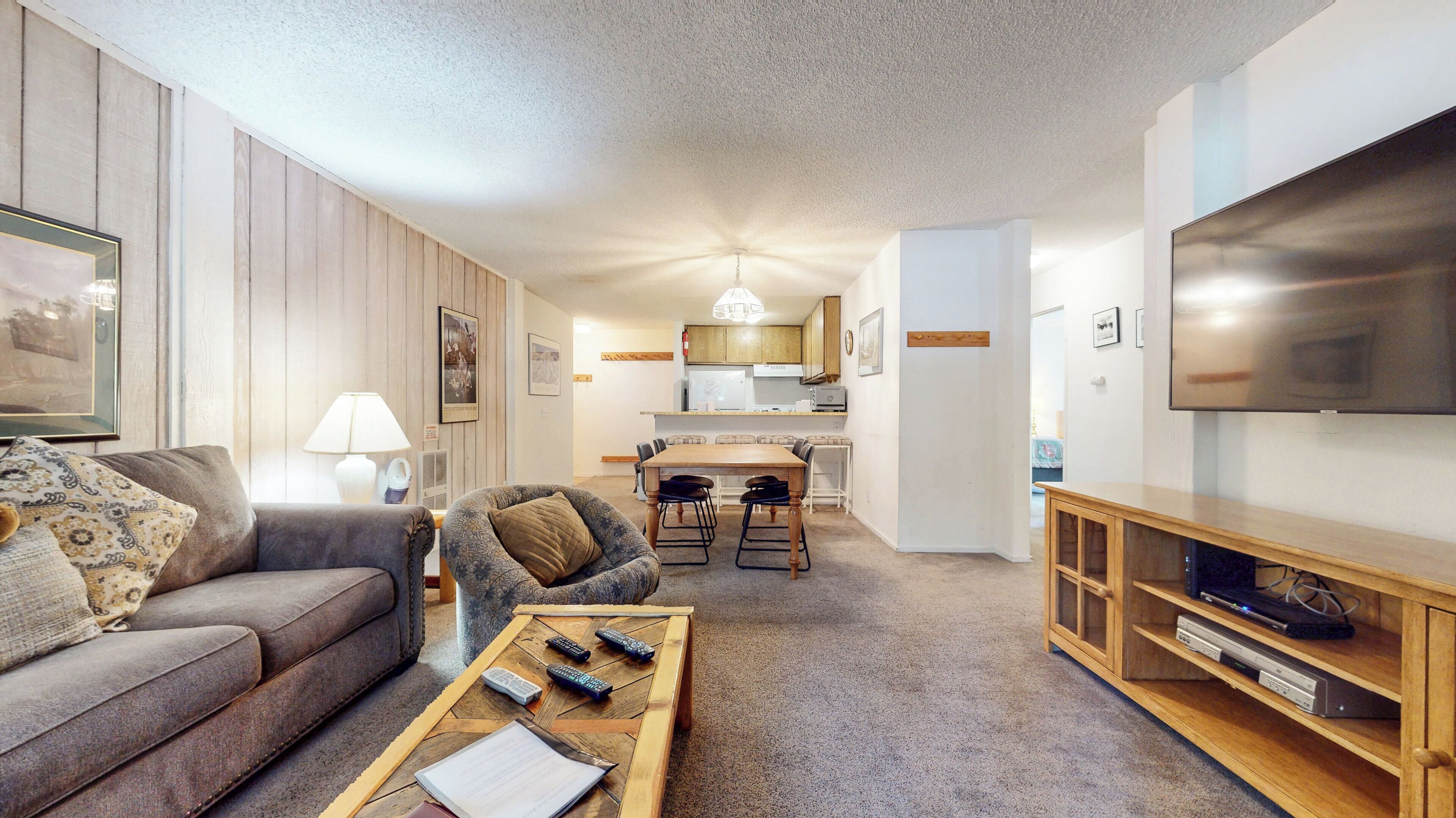 A modern living room with a sofa, a coffee table with remotes, a TV on a wooden stand, and a kitchen in the background.