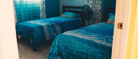 A bedroom with two beds, each with a blue ocean-themed bedspread, a bedside fan, and curtains with a floral pattern.