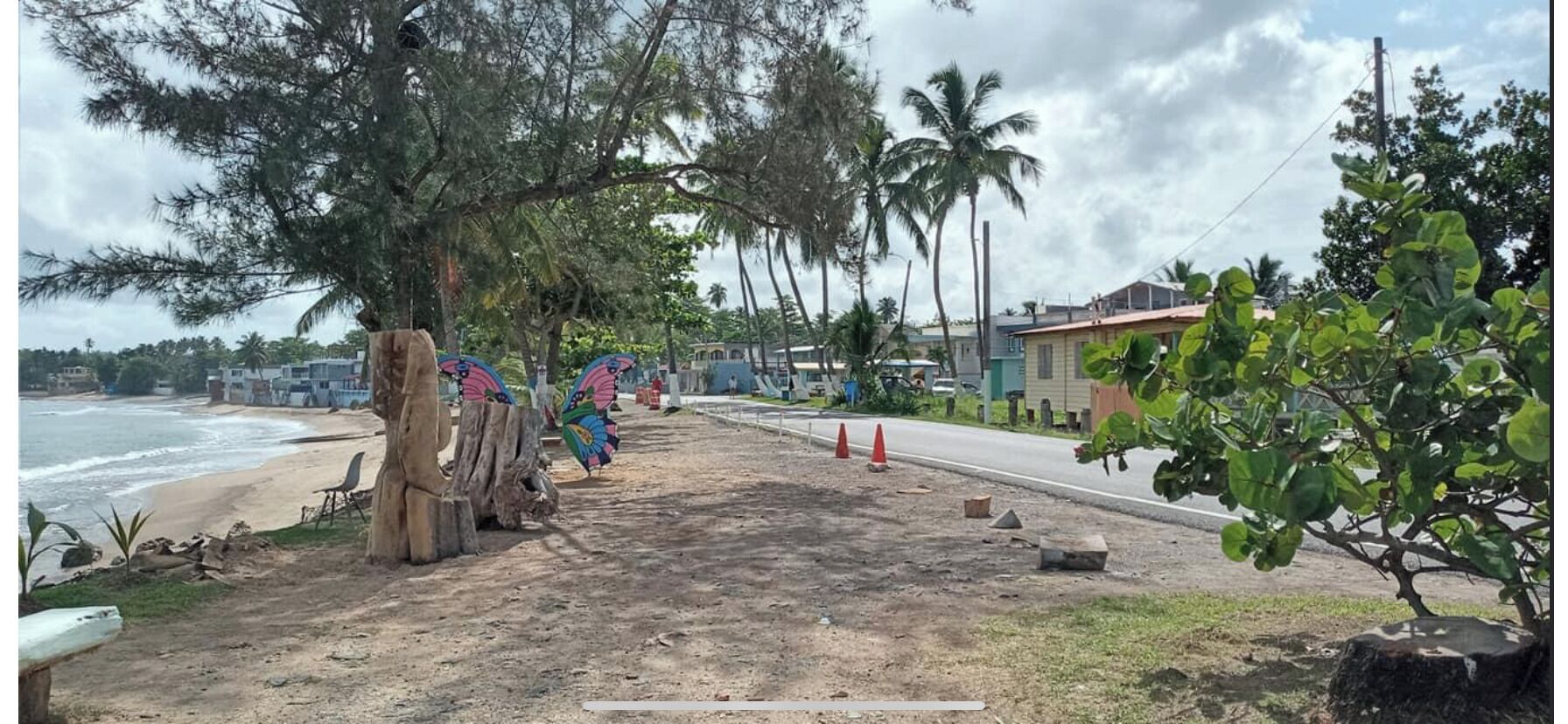 A coastal road with a dirt path, palm trees, and houses in the background.
