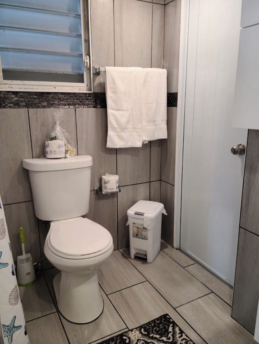 Un baño con un inodoro, un cubo de basura pequeño, una toalla colgada en la pared y una ventana con persianas.