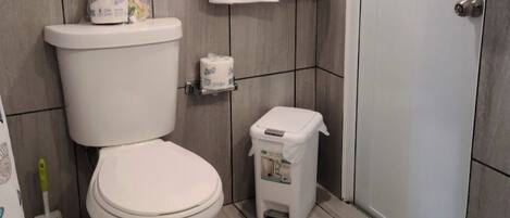 Un baño con un inodoro, un cubo de basura pequeño, una toalla colgada en la pared y una ventana con persianas.