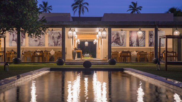 A pool with a reflection, a building with a dining area, and palm trees in the background. - Wirdana Resort & Spa (Boossa)