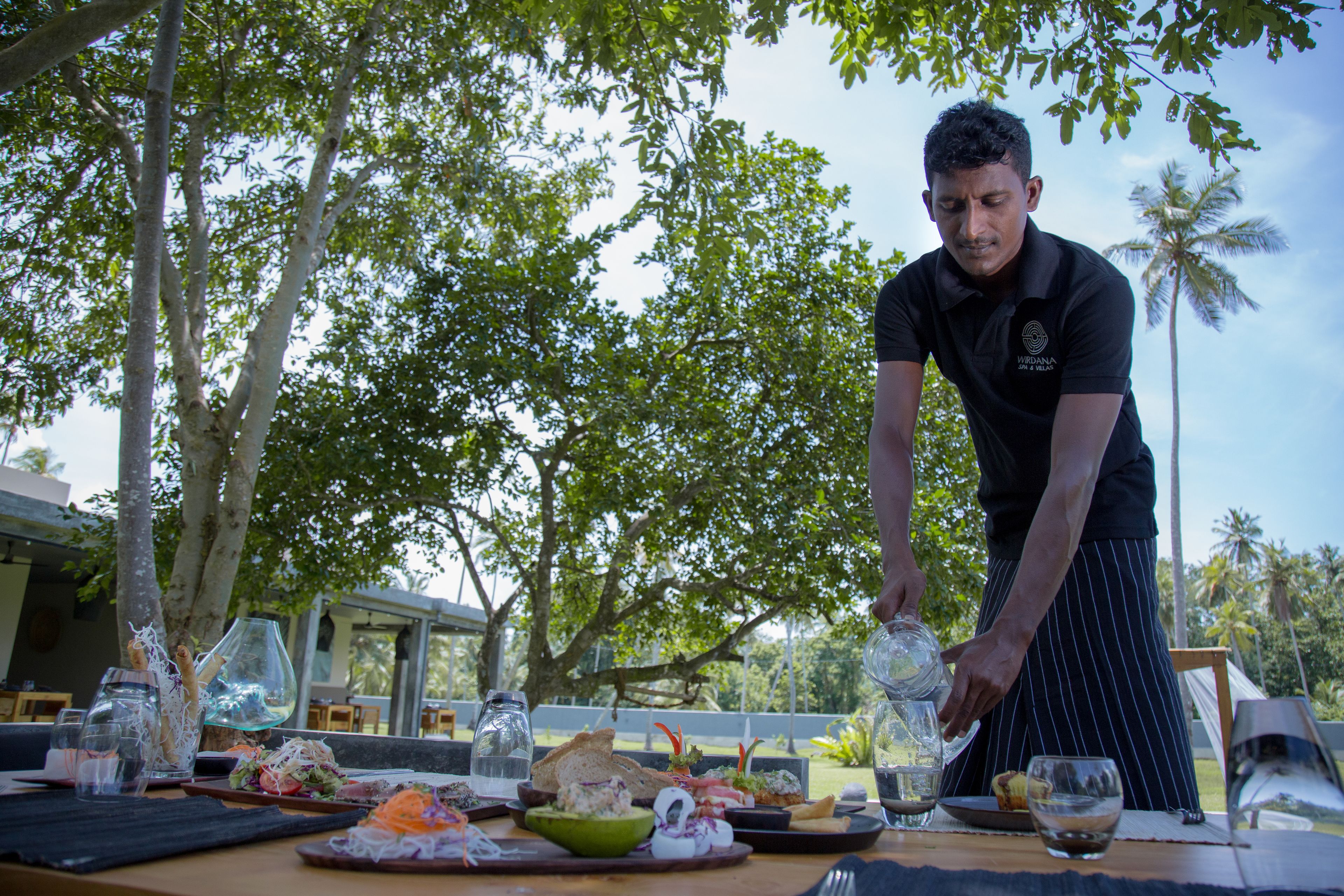 A person serving food at an outdoor dining table with various dishes and drinks.