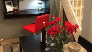 Een eethoek met een tafel voor twee personen, een rode stoel, een vaas met rode bloemen, een fles wijn en een klok aan de muur.