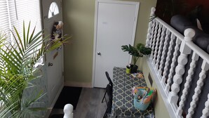 Een kamer met een deur, een klein tafeltje met een tafelkleed met dessin, een bloempot en een witte balustrade met decoratieve details.