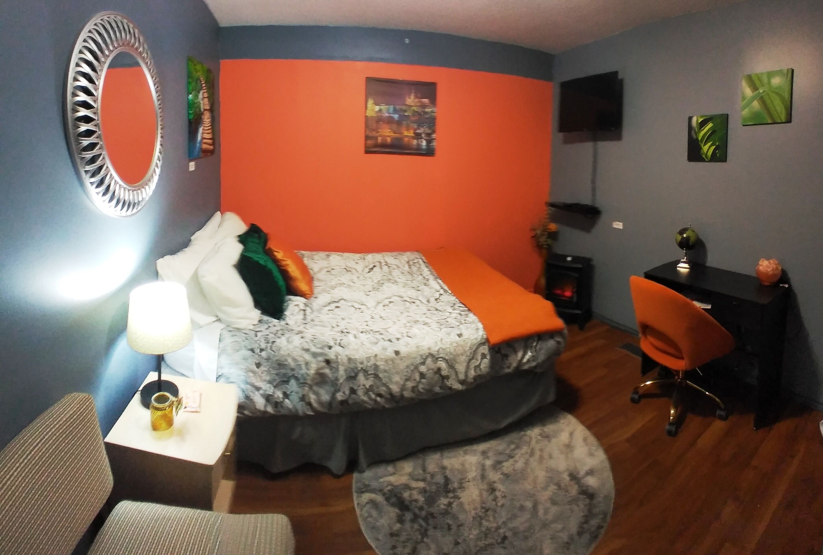 A bedroom with a bed, a desk, an orange chair, a round rug, a mirror, and artwork on the walls.