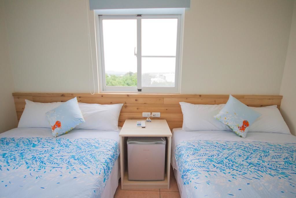 A room with two beds, a small table, a window, and a trash bin.