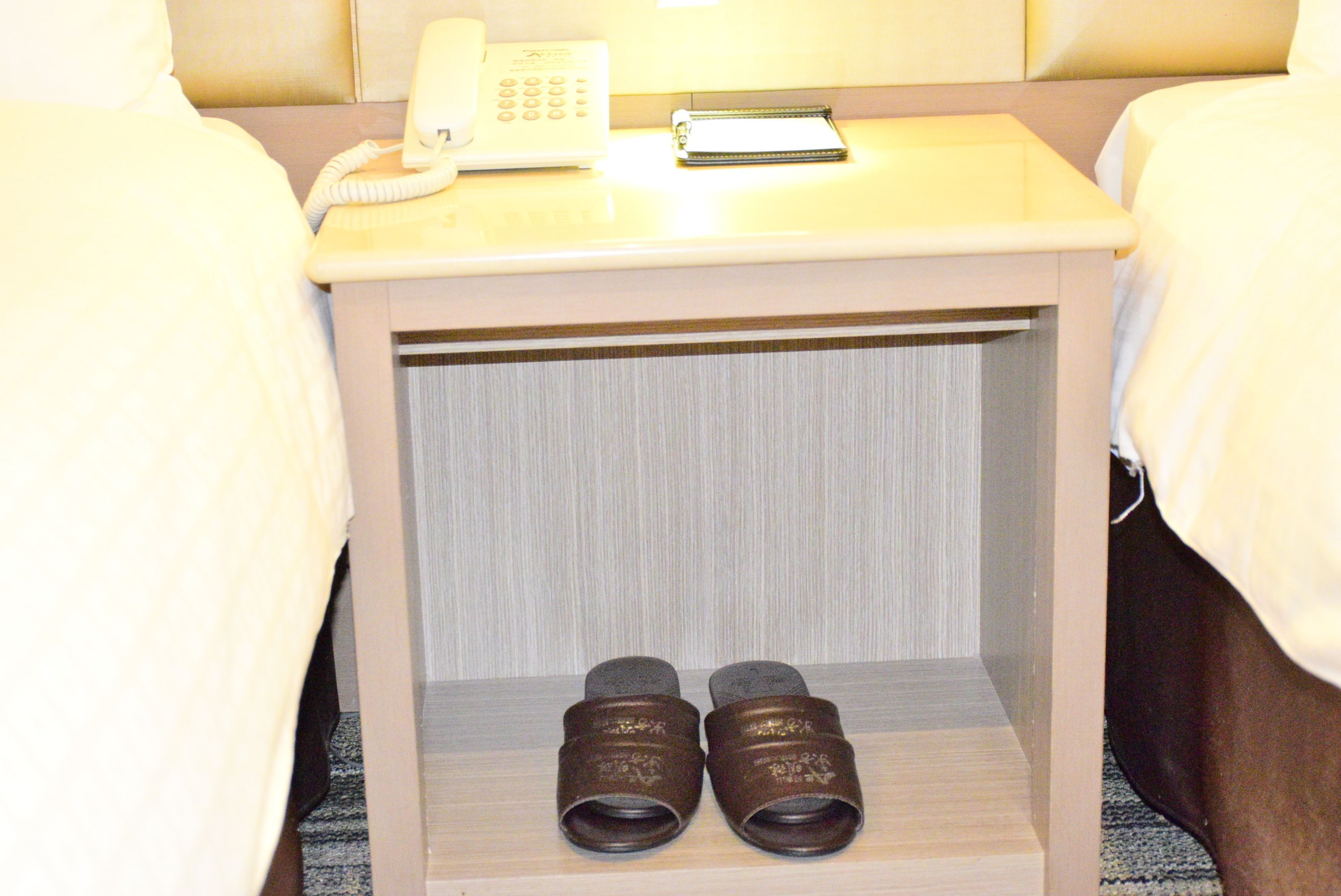 A hotel room with a bedside table, a phone, a notepad, and a pair of shoes on the shelf.