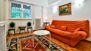 A living room with a patterned rug, a white round table, an orange sofa, and a window with decorative glass. - Groovy Apartment by the Sea with Garden (Alimos)