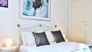 A neatly made bed with white and dark pillows, a bedside table with a lamp, and a framed picture of a lighthouse on the wall. - Groovy Apartment by the Sea with Garden (Alimos)