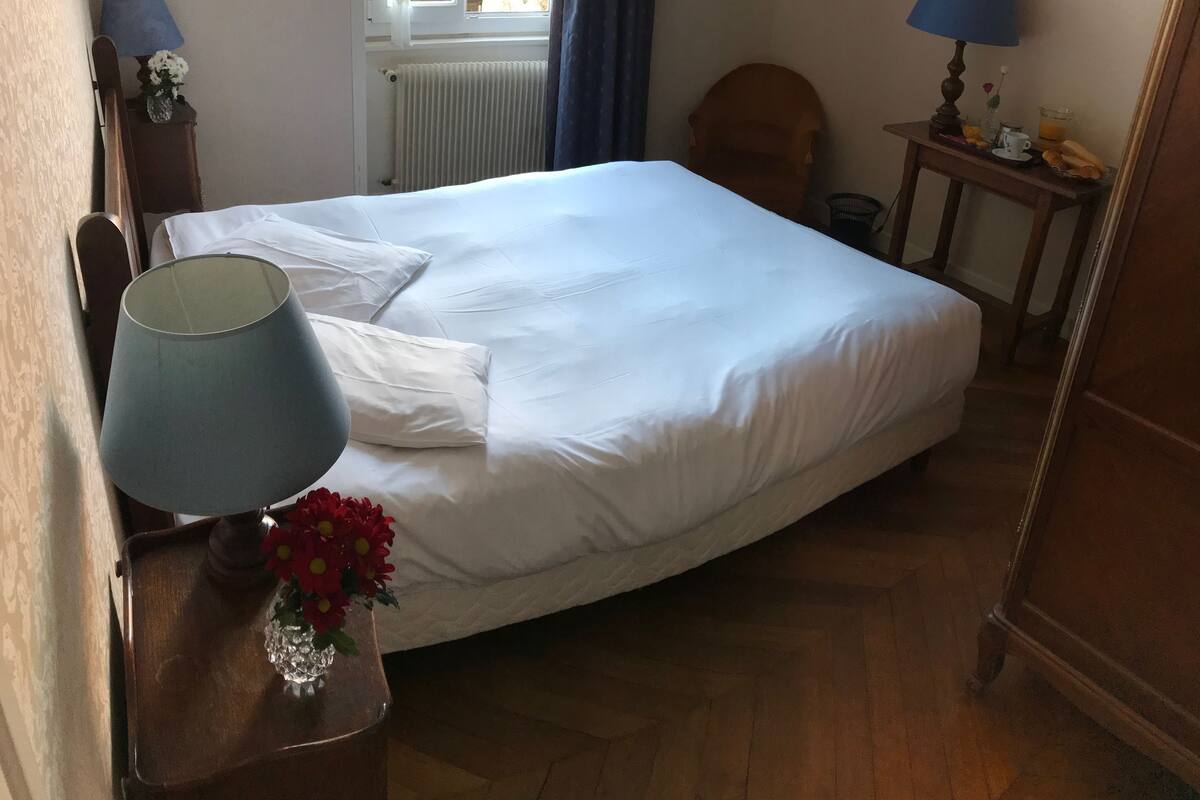 Una cama bien hecha con sábanas blancas, una lámpara de noche, una mesita con un jarrón de flores y una silla.