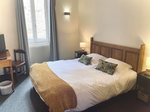 A hotel room with a bed, a desk, a chair, a television, and a window with curtains. - Hôtel des Cymaises (Semur-en-Auxois)