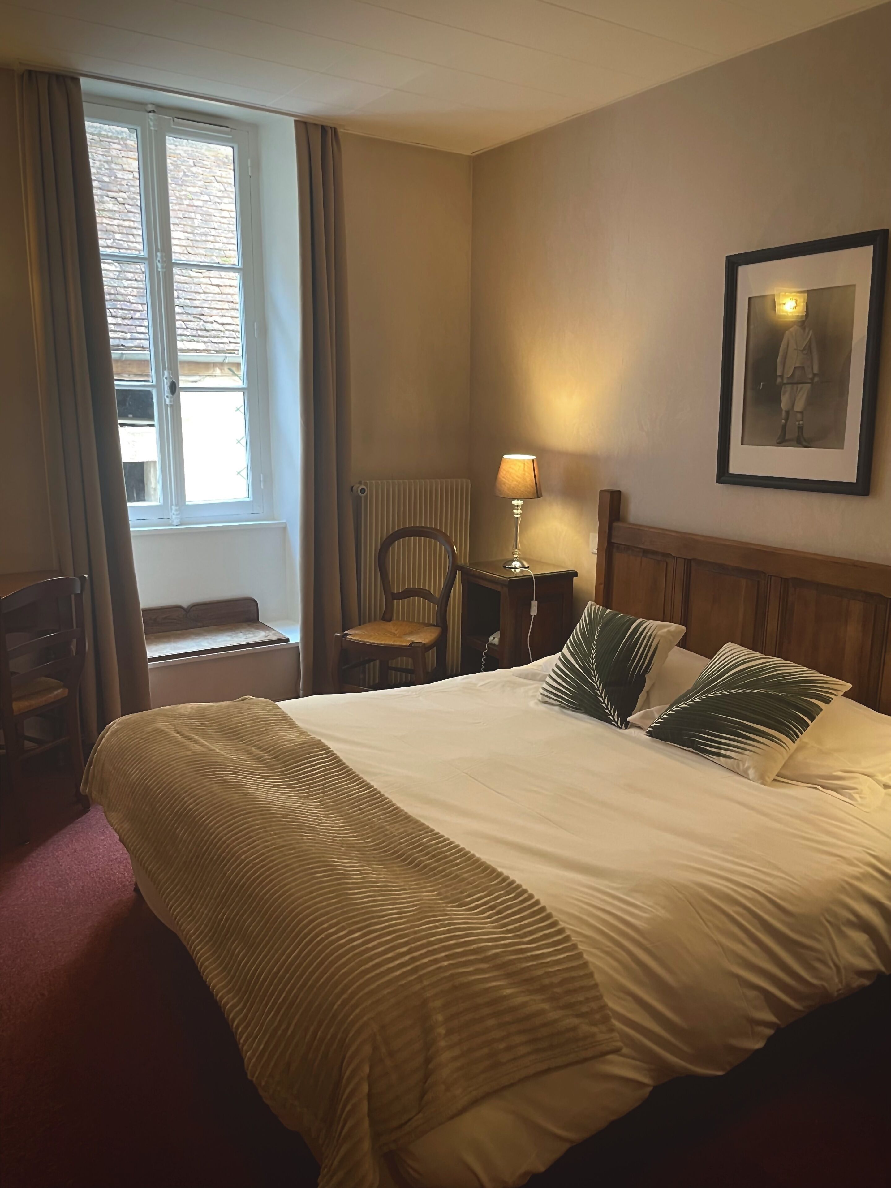A hotel room with a bed, a chair, a table with a lamp, a radiator, and a window with curtains.