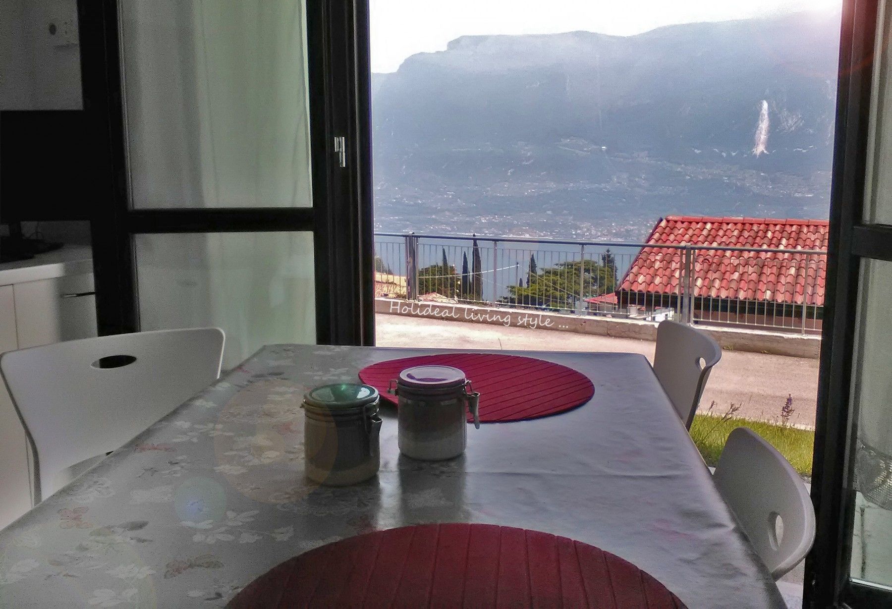 A table with a red placemat, two jars, and a white tablecloth. A window reveals a mountainous landscape and a red-roofed building.