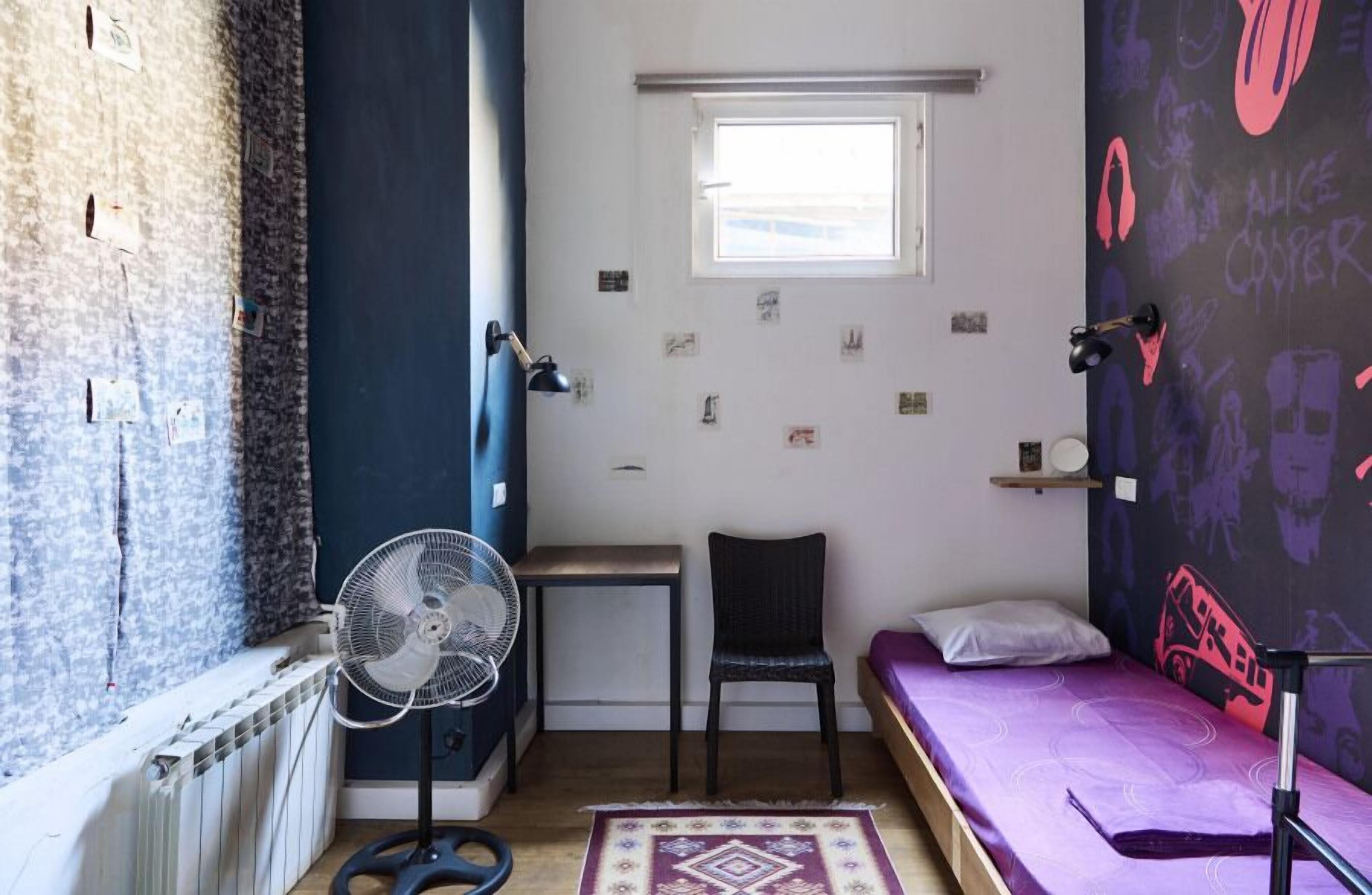 A small, compact room with a bed, a chair, a desk, and a standing fan.