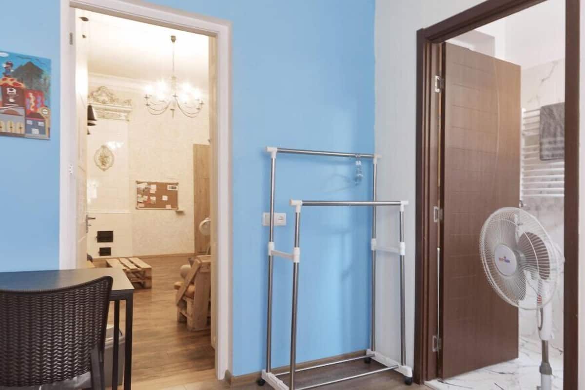 A room with a blue wall, a wooden door, a white fan, a metal rack, and a table with chairs.