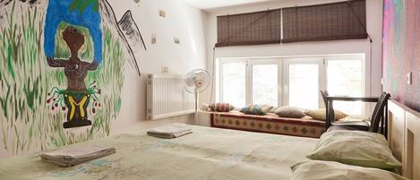 A bedroom with a bed, a radiator, a window with blinds, a chair, and a wall with a mural.