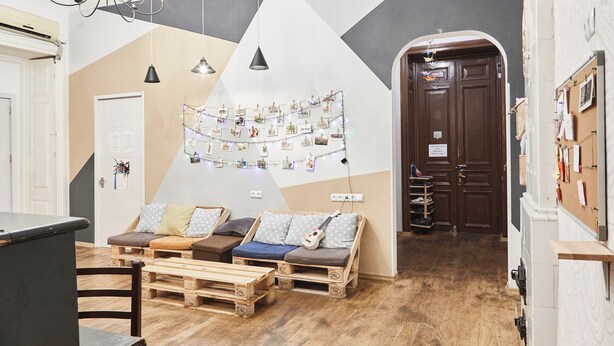 A cozy room with a wooden floor, a sofa, a dining table, and a wall decorated with pictures and lights. - Moosica Hostel (Tbilisi)