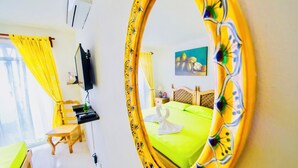 Ein Hotelzimmer mit einem grünen Bett, einem Holzstuhl, einem Schreibtisch, einem Fernseher und einem Spiegel mit floralem Rahmen.