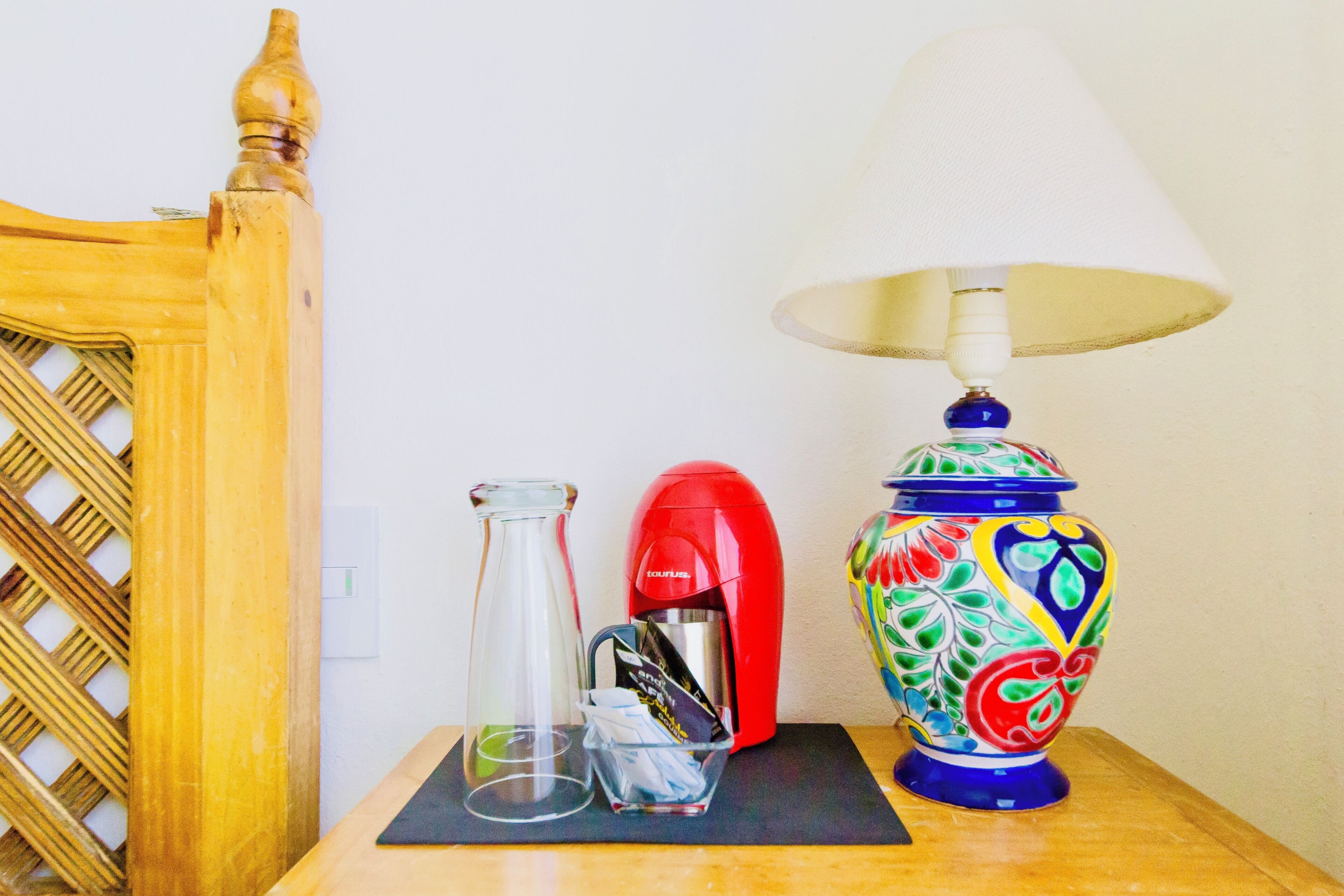 Une chaise en bois, une table avec une lampe, une cafetière rouge, un pichet en verre et un pot en céramique aux couleurs vives.