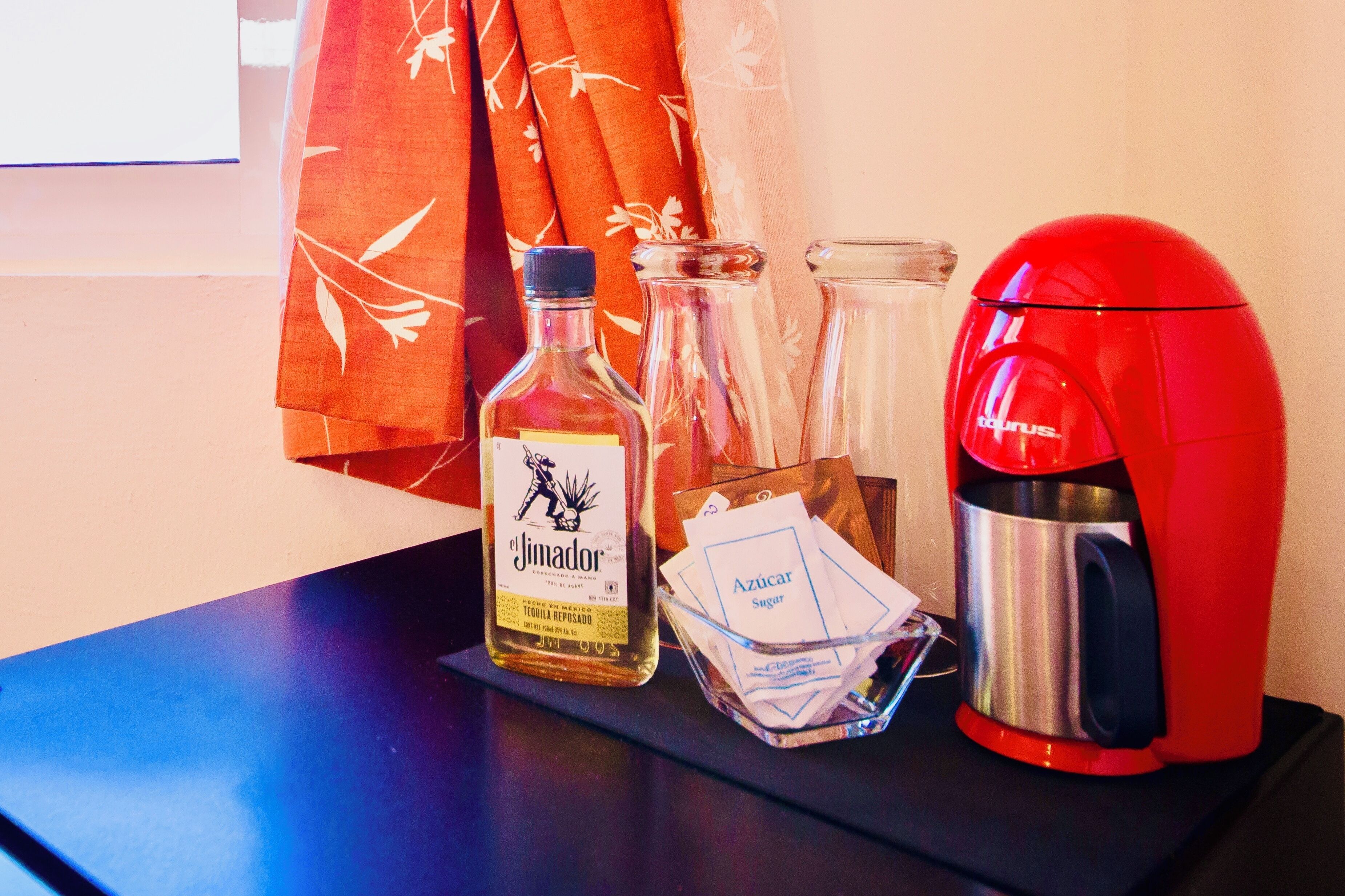 Une table avec une bouteille de tequila Jimador, un pichet en verre, des sachets de sucre et une cafetière rouge.