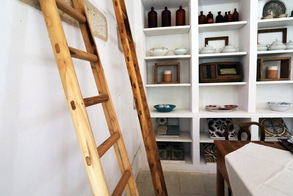 A room with a wooden ladder leaning against a white wall, shelves with various kitchenware, and a table with a white tablecloth.