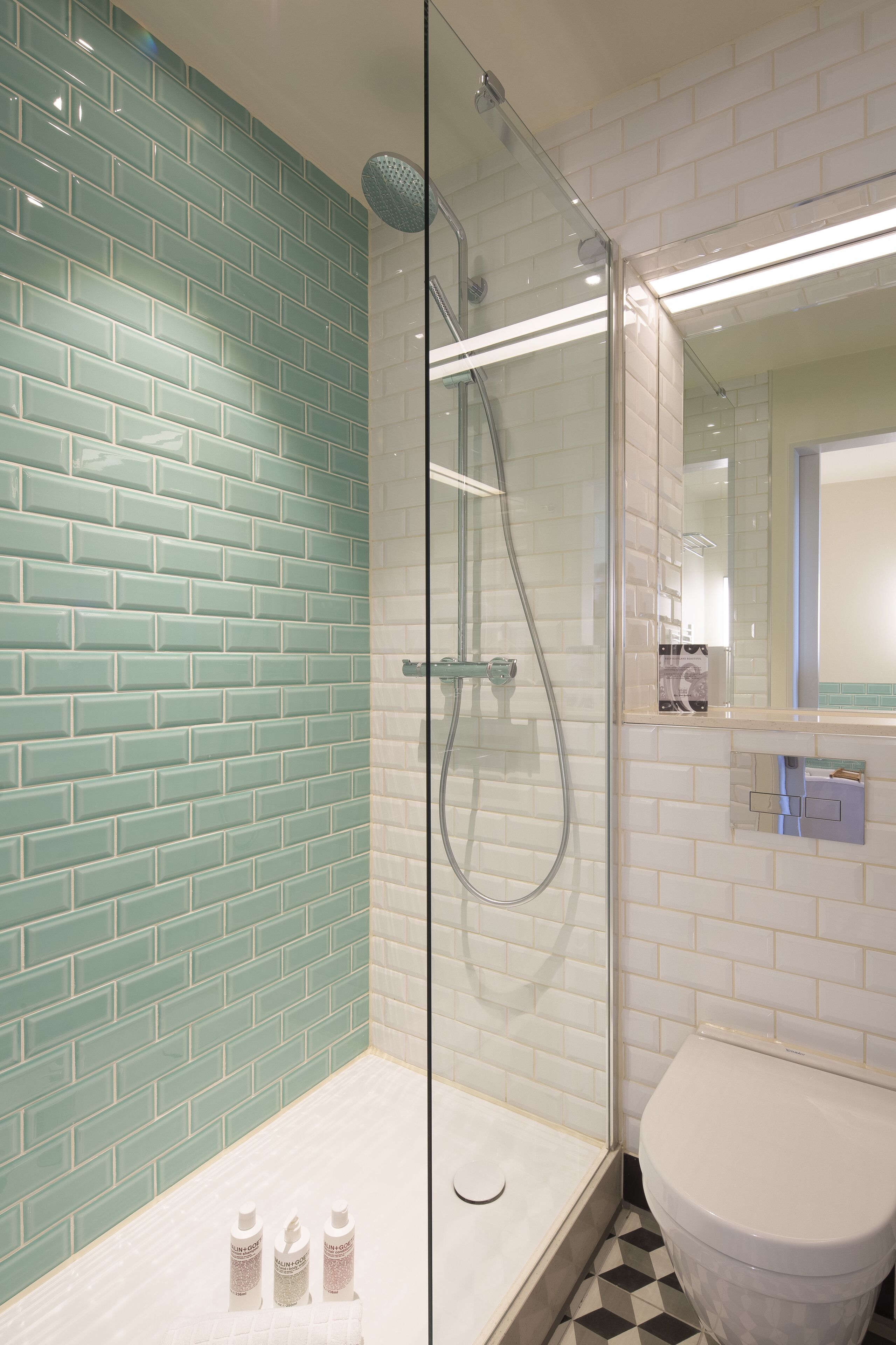 Un baño con ducha envuelta en vidrio, paredes con azulejos blancos y turquesa, un inodoro blanco y un espejo sobre el lavabo.
